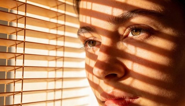 Gazing woman behind wooden blinds casting striped sunlight across face