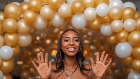 Smiling woman raising hands under gold and white balloon arch at festive party celebration