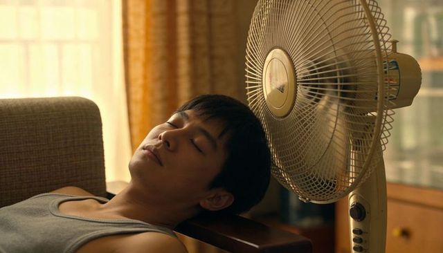 Young asian man resting by pedestal fan in sunlit living room, cooling and relaxing