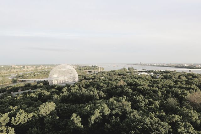 Aerial view of a large, iconic biosphere dome surrounded by dense, verdant forest with a river and cityscape background. Ideal for use in environmental conservation projects, urban development concepts, tourism brochures, and presentations focused on sustainability. Provides a balanced merging of natural beauty and human engineering.