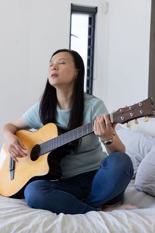 Mature asian woman playing acoustic guitar sitting cross-legged on bed in serene morning