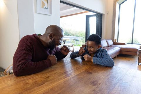 Playful african american father and son mock-boxing at wooden dining table in sunlit home