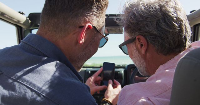 A gay couple is on a summer road trip to the beach and sharing a happy moment while using a smartphone. Great for themes related to LGBT travel, friendship, summer vacations, technology use outdoors, and road trips. Fits well for travel blogs, lifestyle websites, and tourism advertisements.