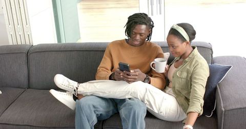 African American Couple Relaxing on Sofa Scrolling Smartphone and Sharing Coffee