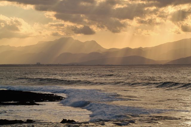 Sun rays filter through clouds over a serene ocean at sunset, casting a golden glow on the water. Mountains in the background add to the tranquil scene. Use this for travel blogs, nature articles, relaxation visuals, or inspirational quotes.