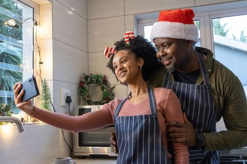 African american couple taking festive kitchen selfie in santa hat and matching aprons