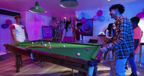 Friends playing pool at neon disco party in games room with disco ball and balloons