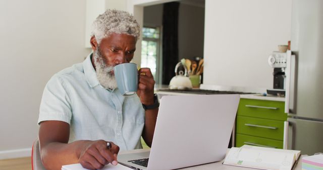 Senior man in casual shirt concentrating on laptop while drinking coffee in a modern kitchen. Ideal for themes around remote work, senior lifestyle, home office, technology use by older adults, and everyday routines.
