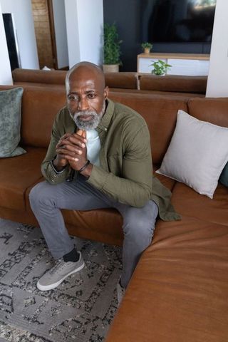 Contemplating african american senior man sitting on brown leather sectional, clasping hands