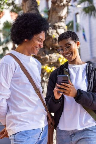 African american couple leaning and smiling while sharing smartphone in sunlit plaza