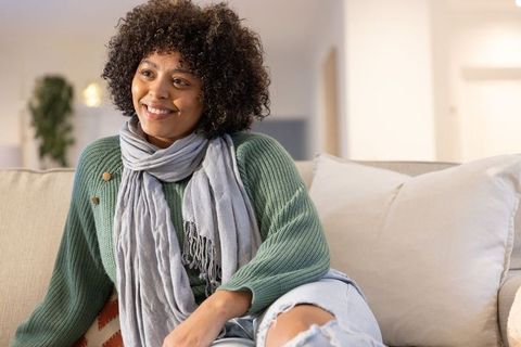 Smiling woman relaxing on sofa wearing green knit sweater and scarf in cozy living room