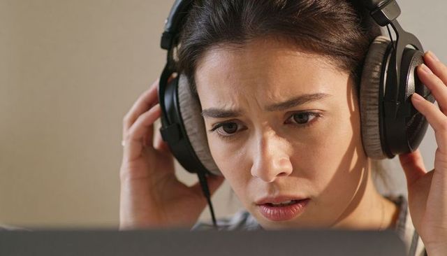 Young woman adjusting over-ear headphones while reacting to unexpected audio on laptop