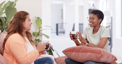 Two friends sharing coffee on sunlit sofa, chatting and laughing in cozy living space