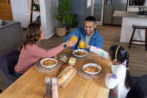Smiling family clinking orange juice glasses over pasta dinner at home kitchen table