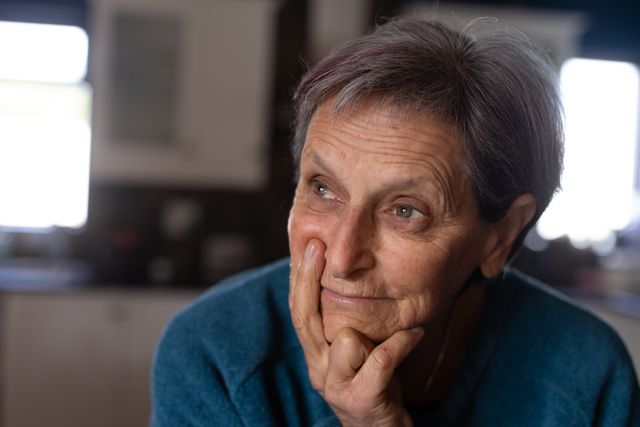 Front view close up of a senior Caucasian woman with short grey hair wearing a blue sweater sitting in her sitting room at home, lining with her chin resting on her hand, relaxing and smiling.