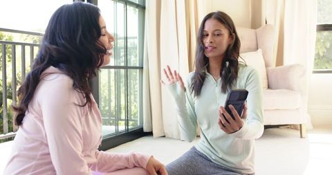 Diverse women chatting in loungewear at home holding smartphone during casual conversation