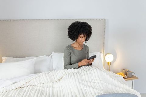 African american woman relaxing in bed using smartphone in cozy neutral bedroom