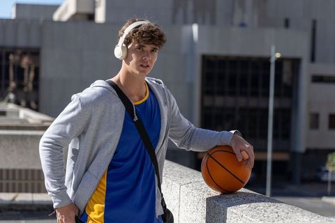Urban teen standing on rooftop listening to music while holding basketball in hoodie