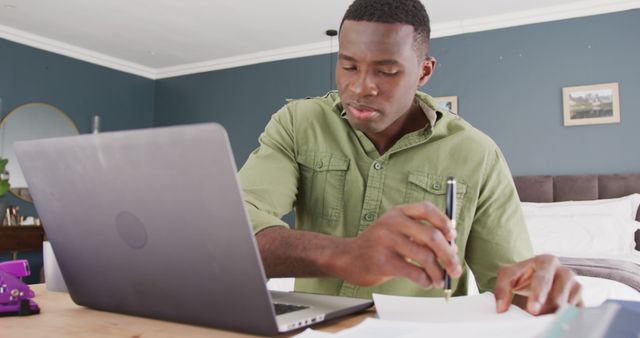 Image of african american man sitting using laptop and writing notes, working in bedroom. Communication, working from home, domestic life, and inclusivity concept.
