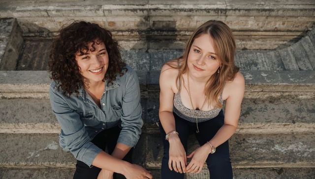Two women sitting on sunlit worn stone steps smiling, wearing denim and strappy top