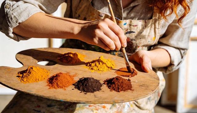 Artist dipping brush into dry earth pigments on wooden palette in sunlit studio close-up