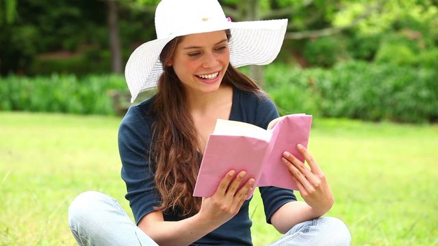 A young woman is sitting on lush green grass in a sunlit park, reading a book and smiling warmly. She wears a white hat and casual clothing, suggesting relaxation and leisure. This image is ideal for content focused on relaxation, reading, book promotions, outdoor activities, lifestyle, and enjoyment of nature. It is suited for media, articles, and blogs related to hobbies, self-care, peaceful living, and personal retreat moments.