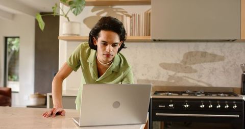 Young man leaning over laptop in modern kitchen wearing pearl necklace and green shirt