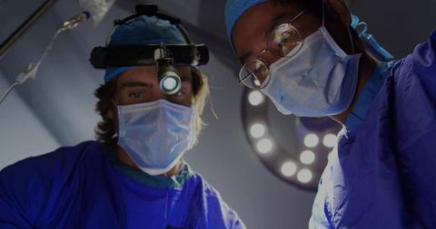 Two Surgeons Performing Surgery Under Overhead LED Surgical Light Wearing Masks and Loupes