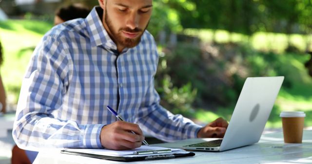 Young businessman sitting in outdoor environment, writing on clipboard and using laptop. Great for advertising remote work, freelance job opportunities, productivity tips in relaxed environments, coffee brand promotions.