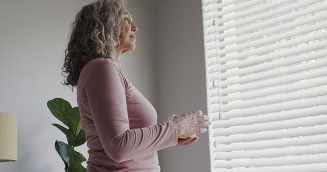 Thoughtful senior caucasian woman holding tea cup and looking out of window at home, copy space. Retirement, contemplation, wellbeing, domestic life and senior lifestyle, unaltered.
