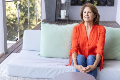 Serene senior woman smiling and sitting on sunlit balcony sofa wearing orange blouse and jeans