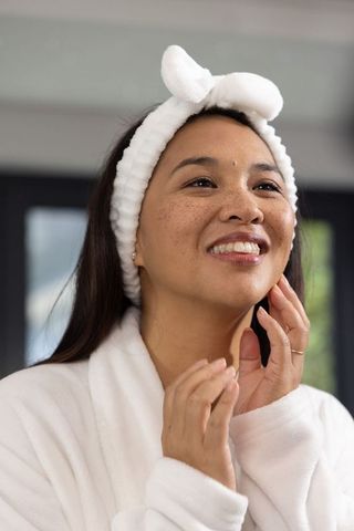 Asian woman smiling and touching glowing face with plush headband and robe at vanity