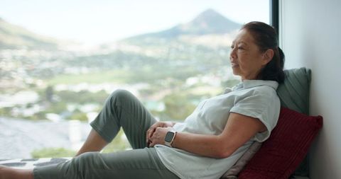 Senior asian woman relaxing on window seat with smartwatch and scenic city view