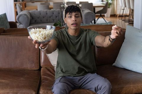 Young man offering popcorn on leather couch watching movie in cozy living room