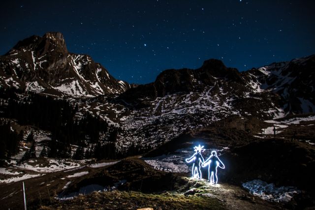 Shows a serene night mountain landscape with light painting of two human figures. Ideal for content related to adventure, stargazing, creativity, and outdoor activities. Perfect for travel blogs, adventure magazines, and night photography inspirations.