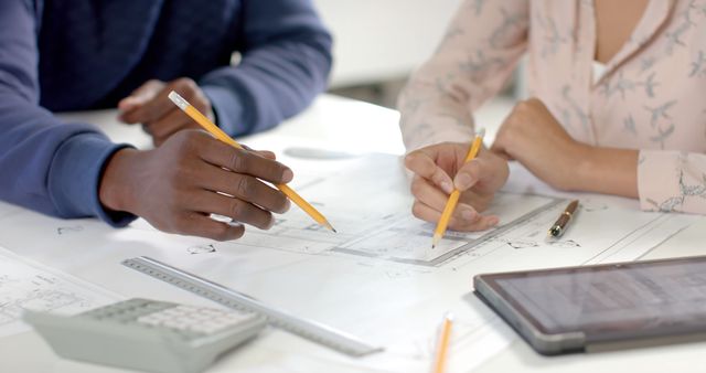 Architects collaborating on blueprint at work desk, discussing project details. Pencil, tablet and calculator aids visualization and measurements. Perfect for content about teamwork, architecture, engineering projects, educational materials, and office dynamics.