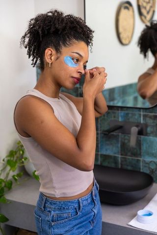 African american woman applying blue under-eye gel patch at bathroom mirror, skincare routine