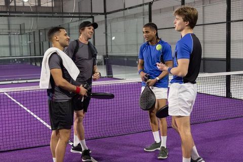 Diverse male teammates chatting at net on purple indoor padel court with paddles