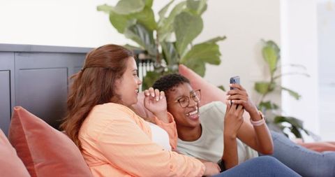 African american friends laughing and scrolling smartphone on cozy sunlit living room sofa
