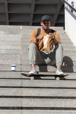 African american man sitting on concrete steps with skateboard holding paper bag lunch