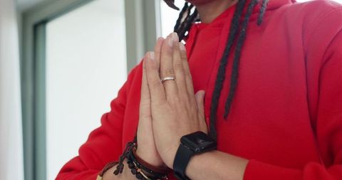Mid-adult man praying by window wearing red hoodie, smartwatch, ring and beaded bracelets