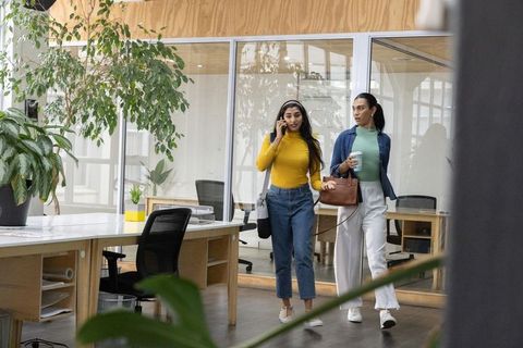 Diverse women coworkers walking through modern office, talking on phone and holding coffee