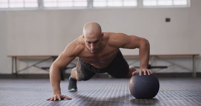 A muscular man is performing push-ups with one hand on a medicine ball in a gym. The scene captures his intense focus and determination during the workout. Use this image for fitness-related content, gym advertisements, workout guides, training programs, and sports health promotions.