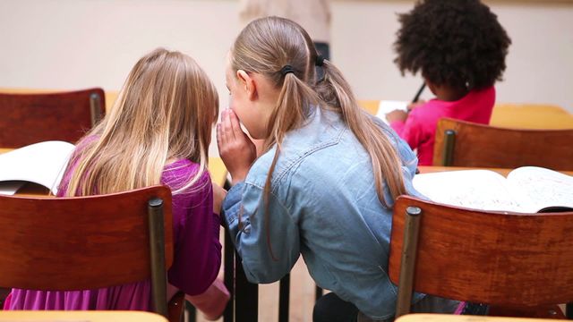 Two young students seen from the back whispering together at their desks in a classroom setting while another student focuses on their work. Ideal for illustrating themes of childhood friendship, secret sharing, education, school life, and student interactions. Perfect for educational materials, children-related articles, or promotional images for schools.