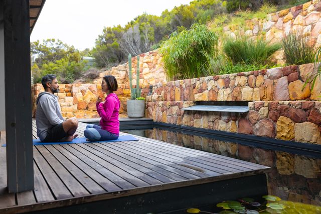 Diverse couple practicing yoga in a serene garden setting, surrounded by natural stone walls and lush greenery. Ideal for use in wellness blogs, fitness websites, and advertisements promoting healthy lifestyles and mindfulness practices.