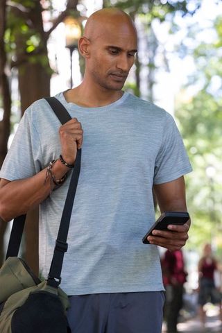 African american man checking phone while carrying olive duffel on tree-lined urban path