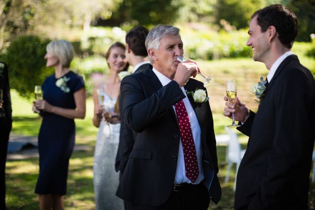 Guests in formal attire are enjoying champagne while attending a wedding in a park. This image can be used for content related to weddings, celebrations, outdoor events, and social gatherings. It is ideal for wedding planners, event organizers, and lifestyle blogs.
