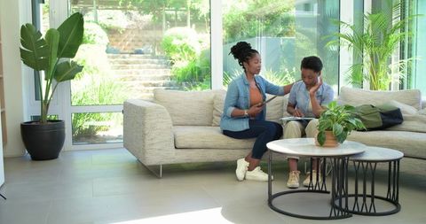 African american mother mentoring teen daughter on sofa using tablet and notebook