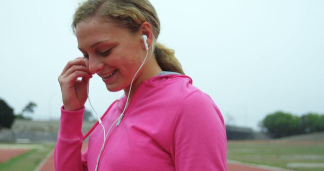 This stock photo features a young woman wearing a pink jacket, enjoying music while exercising outdoors. She is smiling and looks content. This image is ideal for use in fitness and sports advertisements, healthy lifestyle promotions, or athletic wear catalogs.