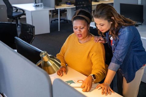 Multiracial female colleagues reviewing data charts and mentoring at open-plan office desk
